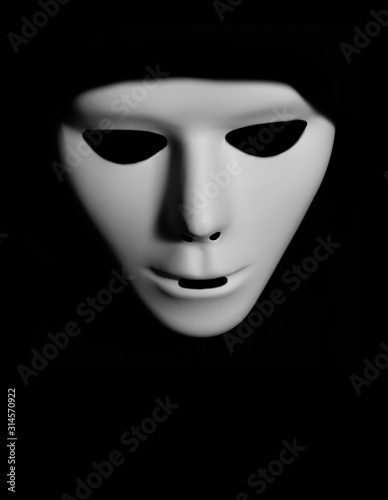 Scary white mask in the dark. Spooky guy in white masquerade. Black, invisible eyes. Black hood. Halloween style. 