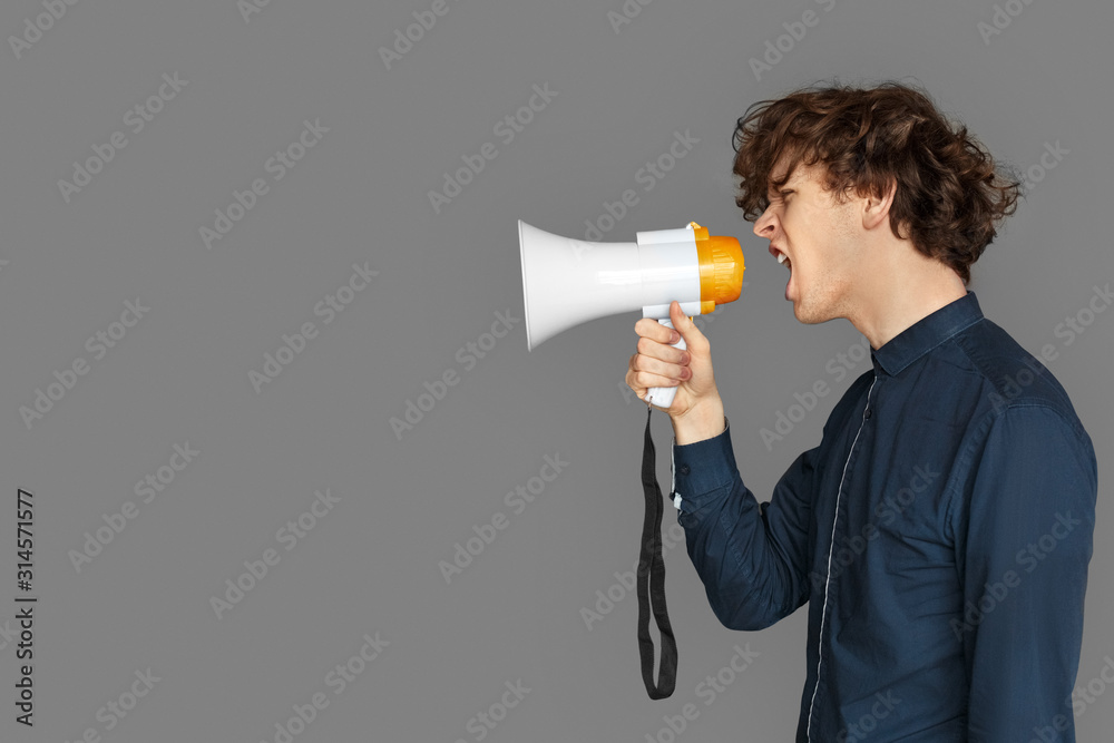 Copy Space for Text or Product. Teenager boy standing isolated on grey shouting emotional at megaphone Copy Space for Text or Product. Teenager boy standing isolated on grey shouting emotional at megaphone