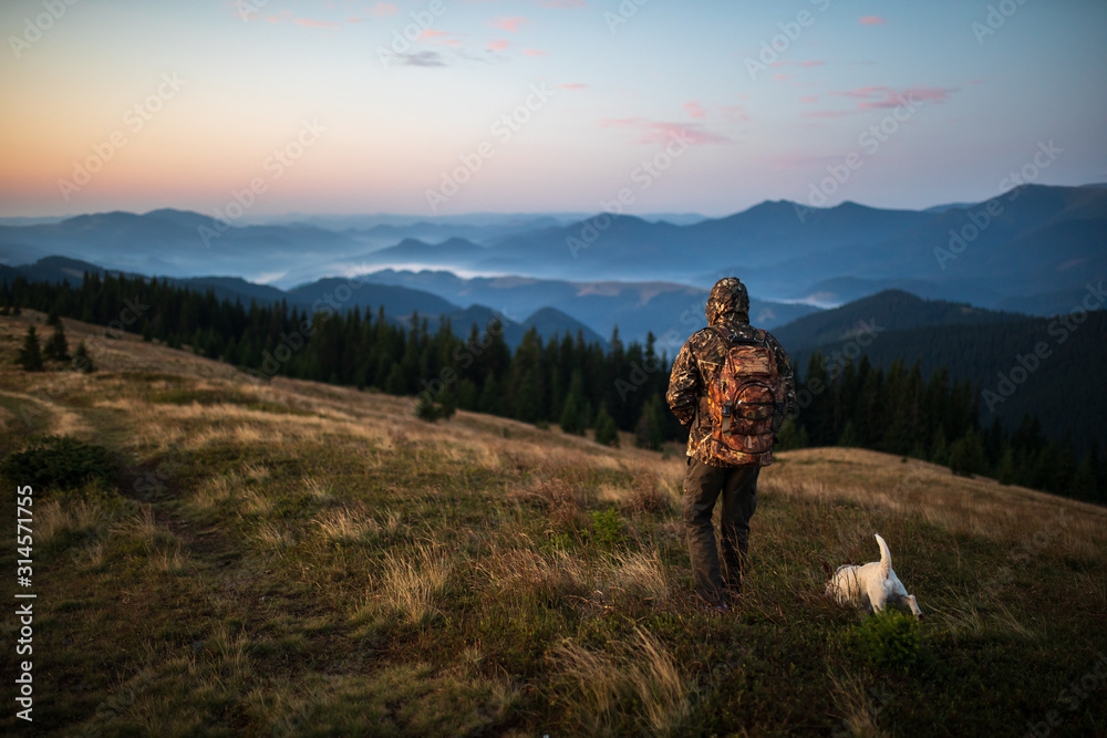 Hunter man dressed in camouflage in the mountains at sunrise. Hunter with dog in the mountains