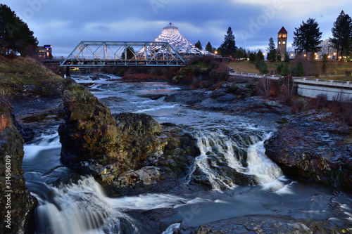 Long exposure, sunrise photo of Spokane Falls on the Spokane River looking over Riverfront Park in downtown Spokane, Washington with a clock tower, bridge and pavilion in the background