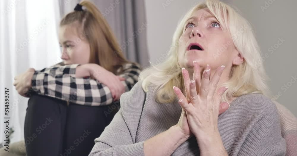 Close-up of worried Caucasian grandmother looking away and up and shaking hands. Upset teenage granddaughter sitting at the background. Conflict of generations. Cinema 4k ProRes HQ. Close-up of worried Caucasian grandmother looking away and up and shaking hands. Upset teenage granddaughter sitting at the background. Conflict of generations. Cinema 4k ProRes HQ.