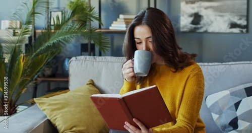 Caucasian young good looking wise woman reading a book and sipping hot tea or coffee while sitting on the couch in the living room.