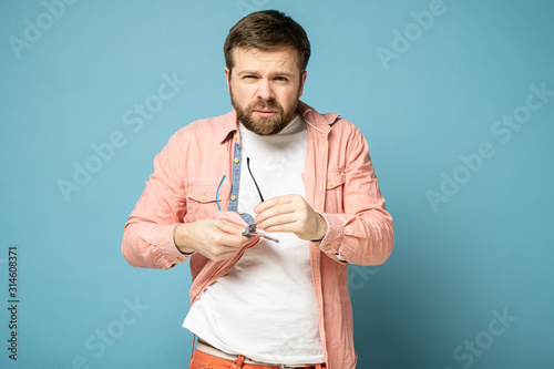 Fényképezés Bearded man cleans glasses, wiping them with a shirt and squinting, looking confidently into the camera