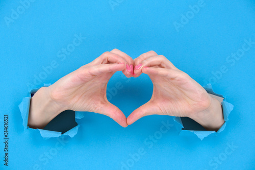 Female hands show heart symbol through ragged holes of a blue torn paper background. Creativity  a copy of space. Concept of love  Valentines day  mother day  social advertising concept.