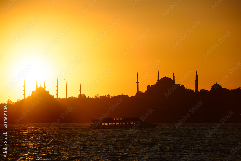 Sun setting over Blue Mosque with Hagia Sophia in silhouette on the Bosphorus with boat Istanbul