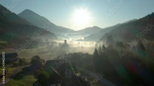 Flying over the misty landscape of the Zell Pfarre Village and Valley in Austria with the mountains in the background and sun rays penetrating through the mist - Aerial shot photo