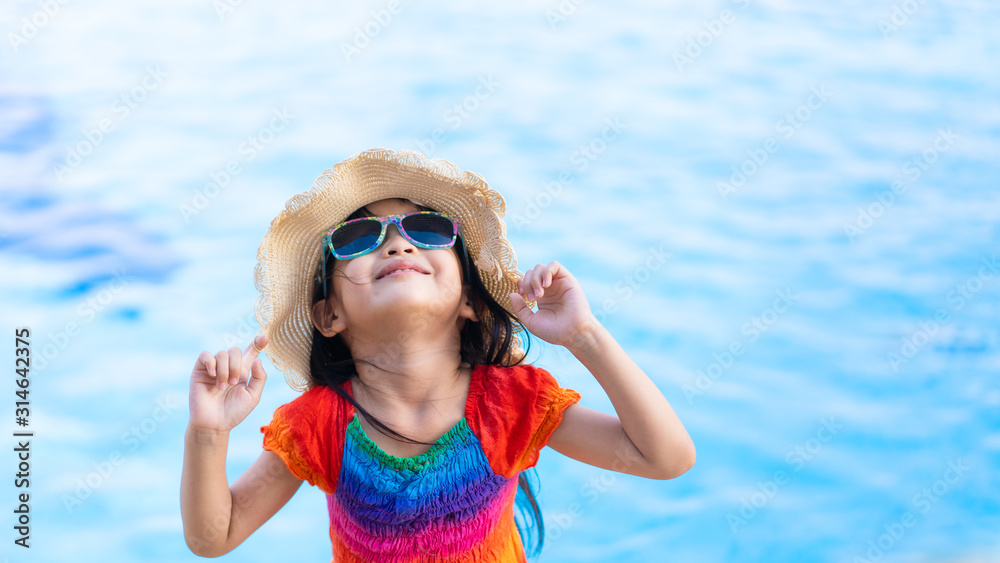 Beautiful asian little girl wear a colorful dress, sunglasses standing near the pool and looking up to the sky, Concept of happy time of summer vacation holiday for kid and family.