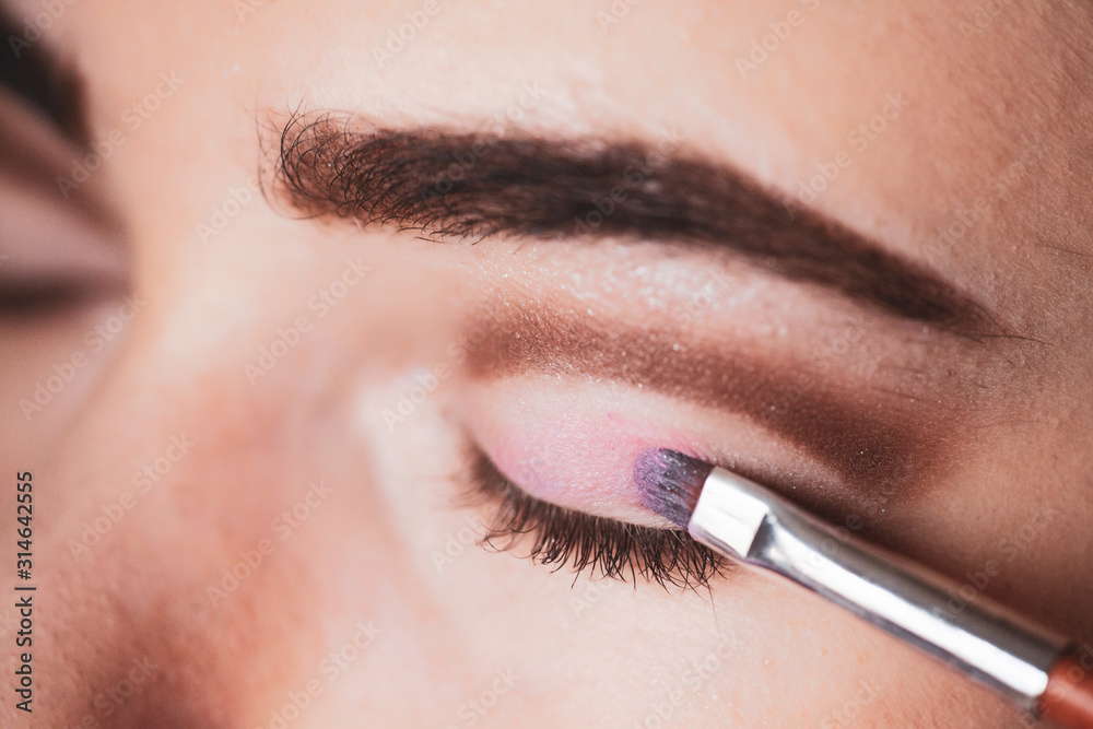 The work of a professional makeup artist. Eyeshadow applying, makeup for eyes closeup. Young woman applies pink colored eyeshadow with make up brush.