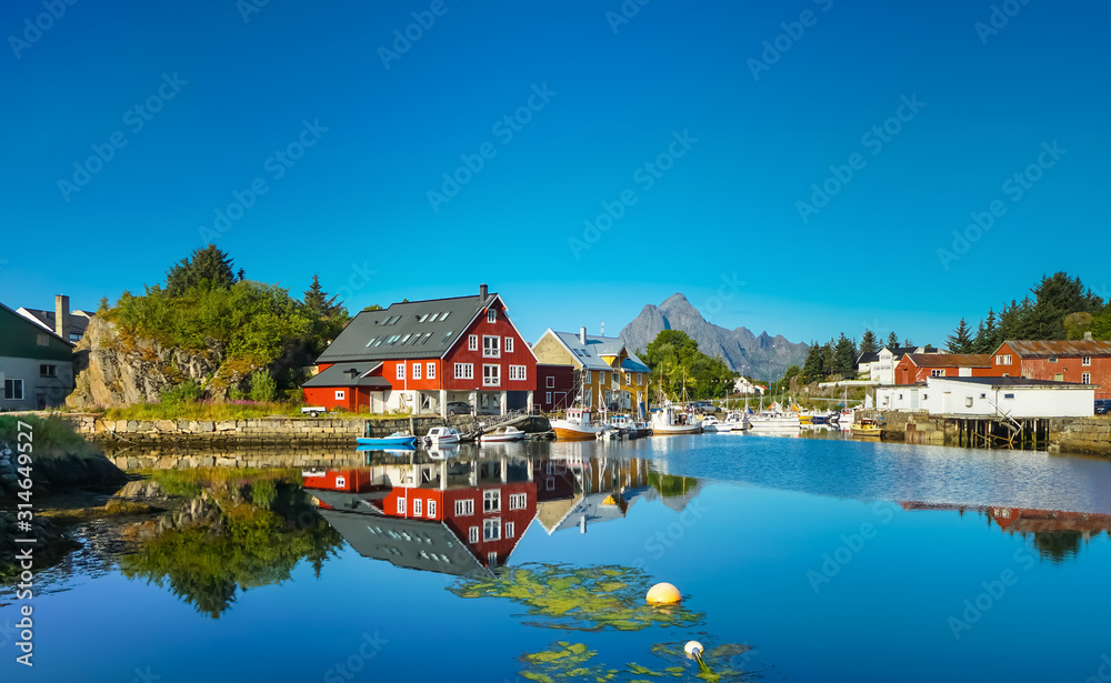 Village on the shore of the fjord, Lofoten Islands, Norway. Sunny day, clear blue sky, colorful house, reflection in the water, forest and mountains in the background. - obrazy, fototapety, plakaty 