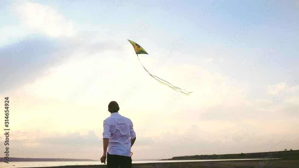 Young man in white shirt flies colorful kite in sky with light clouds at sunset backside low angle shot slow motion