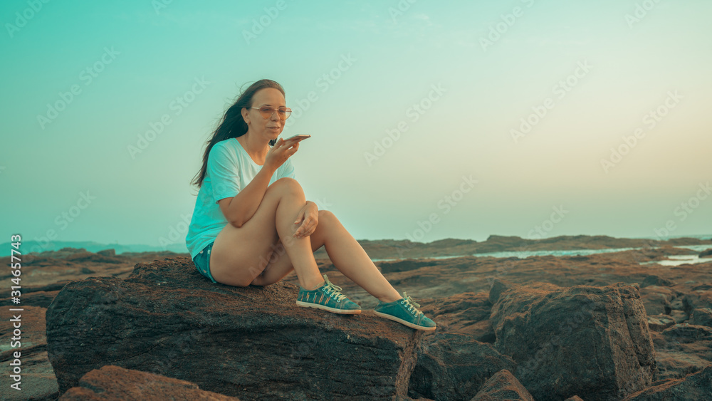 Relaxed woman sitting on stone and recording audio message on smartphone on shore. Side view of pleasant girl spending time enjoying vacation and talking on mobile phone on stony shore. Relaxed woman sitting on stone and recording audio message on smartphone on shore. Side view of pleasant girl spending time enjoying vacation and talking on mobile phone on stony shore.
