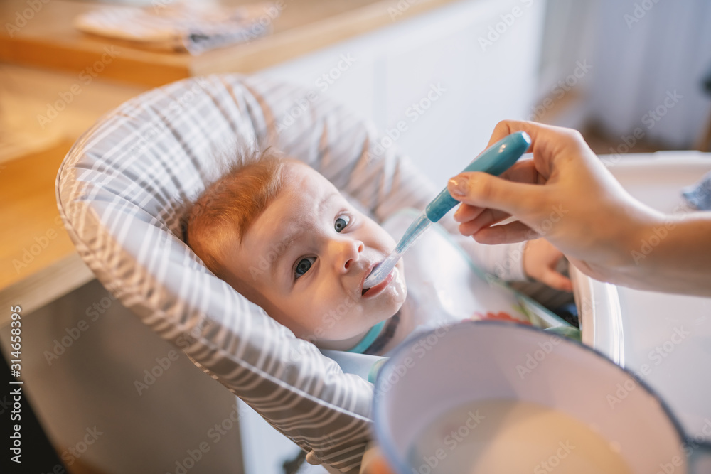 Hungry little baby boy with bib sitting in his chair and taking spoon from his mother. What a delicious food.