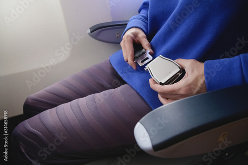Close-up of a young woman fastens a seat belt while sitting in a passenger airplane chair by the window. The concept of safety measures for passenger flights in aircraft