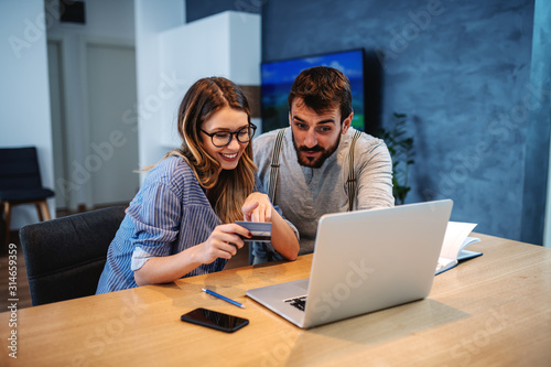 Young handsome caucasian bearded smiling man talking about things they want to buy online. Woman holding and pointing at credit card. On dining table is laptop.