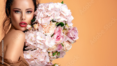 Fototapeta Naklejka Na Ścianę i Meble -  Beautiful white girl with flowers. Stunning brunette girl with big bouquet flowers of roses. Closeup face of young beautiful woman with a healthy clean skin. Pretty woman with bright makeup