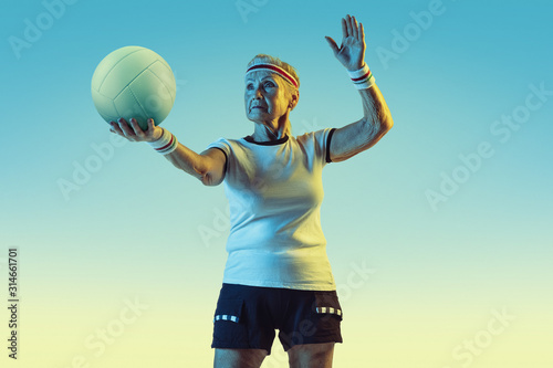 Senior woman in sportwear training in volleyball on gradient background, neon light. Female model in great shape stays active. Concept of sport, activity, movement, wellbeing, confidence. Copyspace.