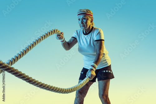 Senior woman in sportwear training with ropes on gradient background, neon light. Female model in great shape stays active. Concept of sport, activity, movement, wellbeing, confidence. Copyspace.