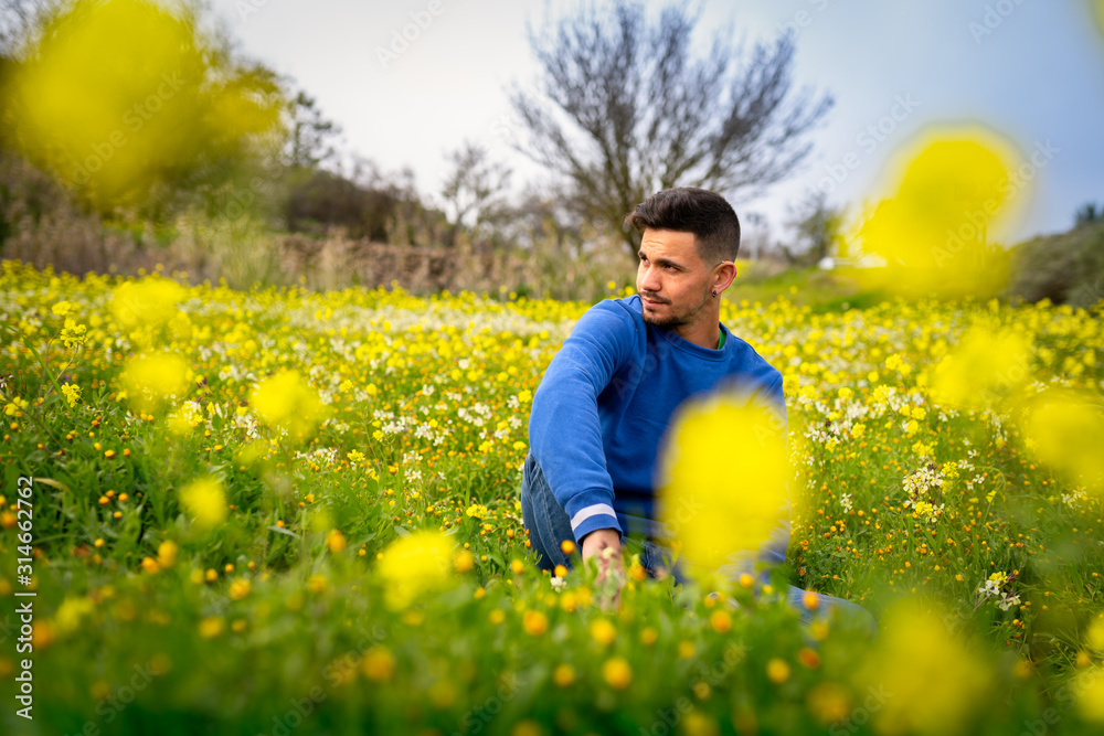 Joven sentado en un campo de flores silvestres, parece oculto detrás de flores amarillas borrosas