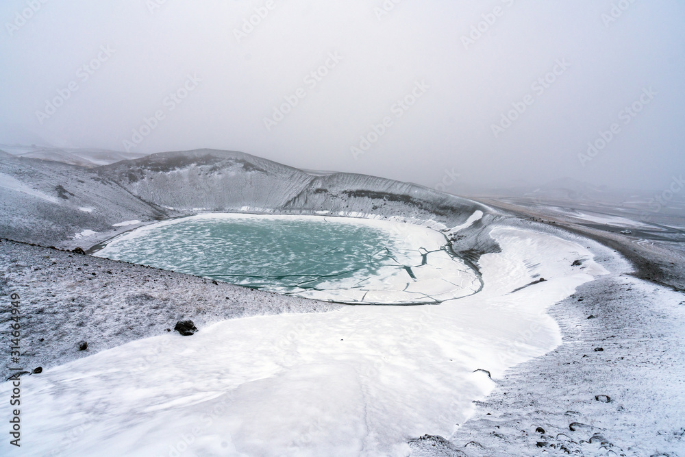 Obraz premium Ovelooking ice covered Viti volcanic crater near Krafla geothermal area in Iceland. Foggy and snow covered landscape with frozen green lake at the bottom. Volcano, energy and weather concept.