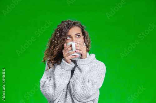 Young beautiful woman standing with cup of hot coffee wearing knitted warm sweater, dreaming about something inspiring.