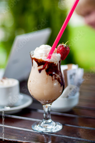 A glass of cocktail milkshake frappe decorated with strawberry and chocklate on the wooden table with laptot and a cup of coffee on background. Vacation concept photo
