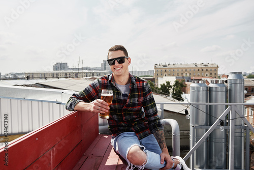 Happy man in eyewear drinking beer at bar or pub on roof, copy space