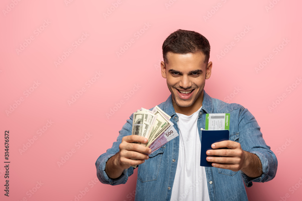 Man holding passport with boarding pass and cash while smiling at camera on pink background Man holding passport with boarding pass and cash while smiling at camera on pink background