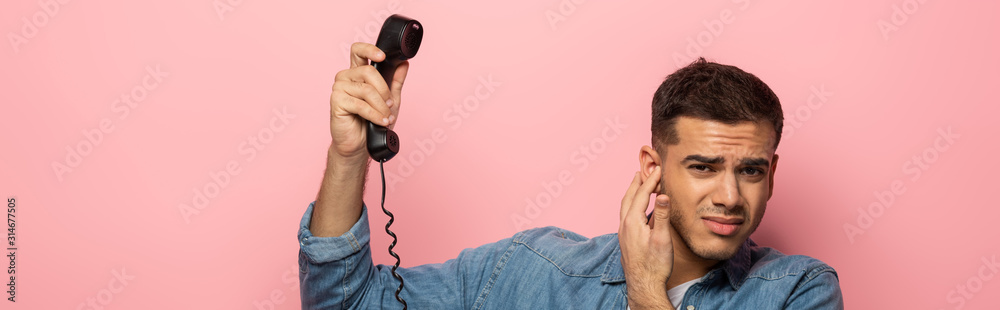 Panoramic shot of confused man holding telephone handset and closing ear isolated on pink Panoramic shot of confused man holding telephone handset and closing ear isolated on pink