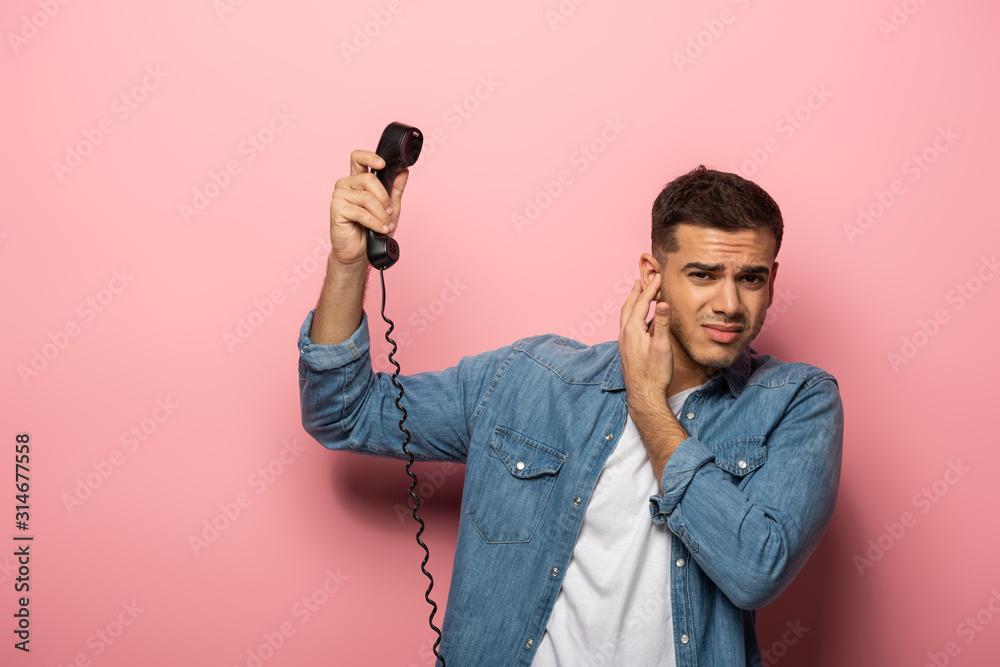 Confused man with telephone handset closing ear on pink background Confused man with telephone handset closing ear on pink background