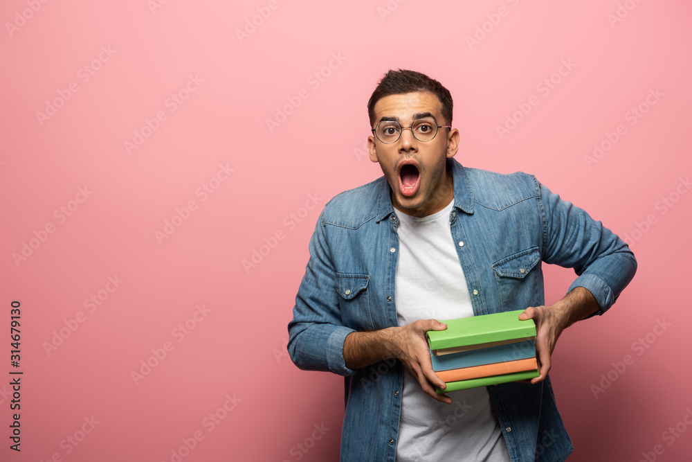 Shocked man holding books and looking at camera on pink background Shocked man holding books and looking at camera on pink background