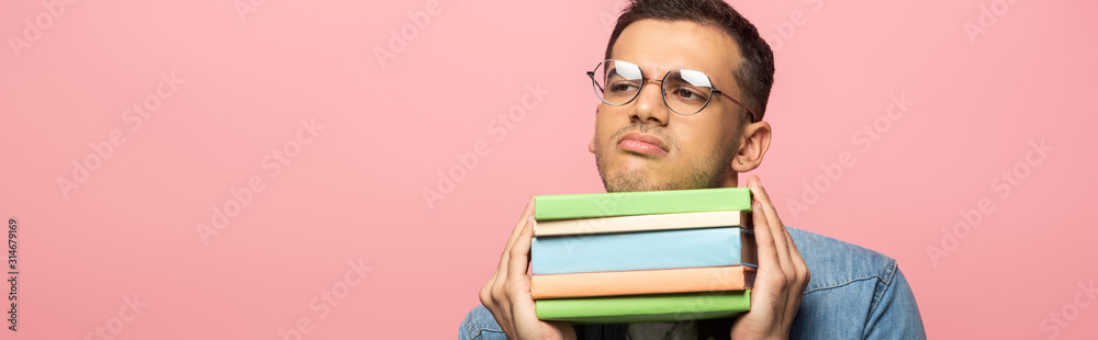 Skeptical man with books looking away isolated on pink, panoramic shot Skeptical man with books looking away isolated on pink, panoramic shot