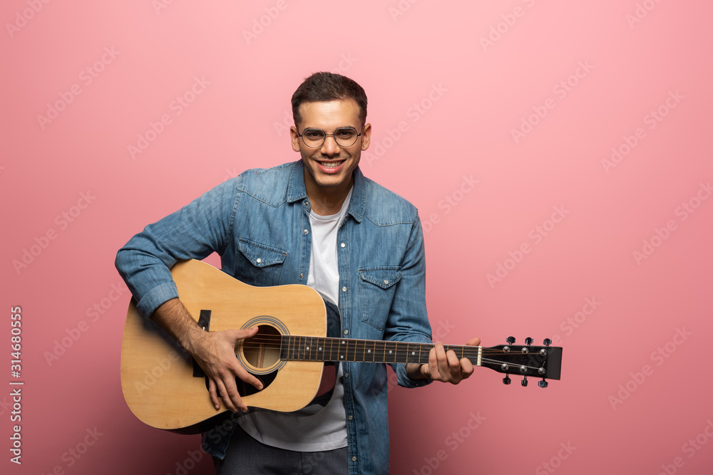 Obraz premium Young man smiling at camera and playing acoustic guitar on pink background