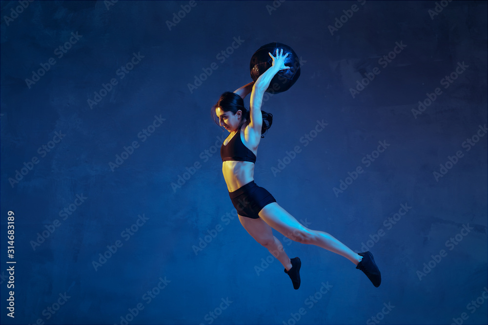 Caucasian young female athlete practicing on blue studio background in neon light. Sportive model in flight, jump with ball, training. Body building, healthy lifestyle, beauty and action concept.
