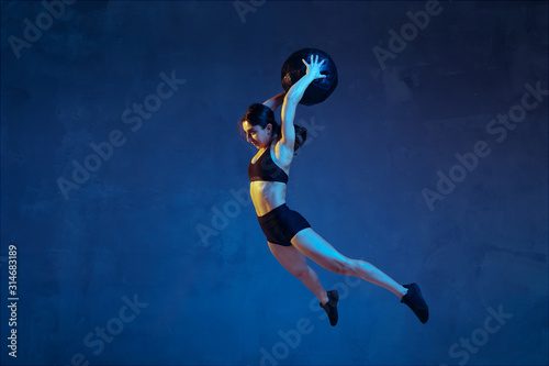 Caucasian young female athlete practicing on blue studio background in neon light. Sportive model in flight  jump with ball  training. Body building  healthy lifestyle  beauty and action concept.