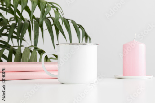 Enamel mug mockup with books and a candle on a white table and a palm plant.