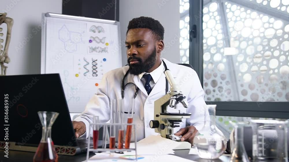 Attractive portrait of serious 35-aged confident black-skinned medical assistant which looking into microscope and writing down the results on the laptop and papers Attractive portrait of serious 35-aged confident black-skinned medical assistant which looking into microscope and writing down the results on the laptop and papers
