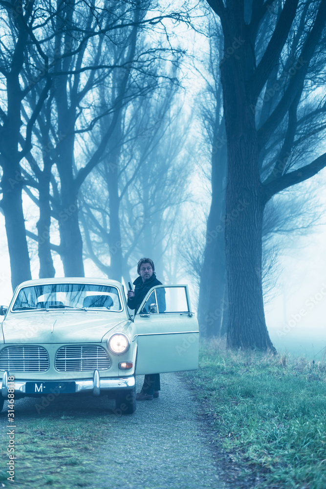 Man with pistol in black jacket standing behind door of classic car during misty dusk. Man with pistol in black jacket standing behind door of classic car during misty dusk.