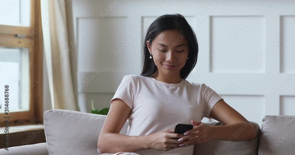 Smiling millennial asian woman using smart phone app on sofa