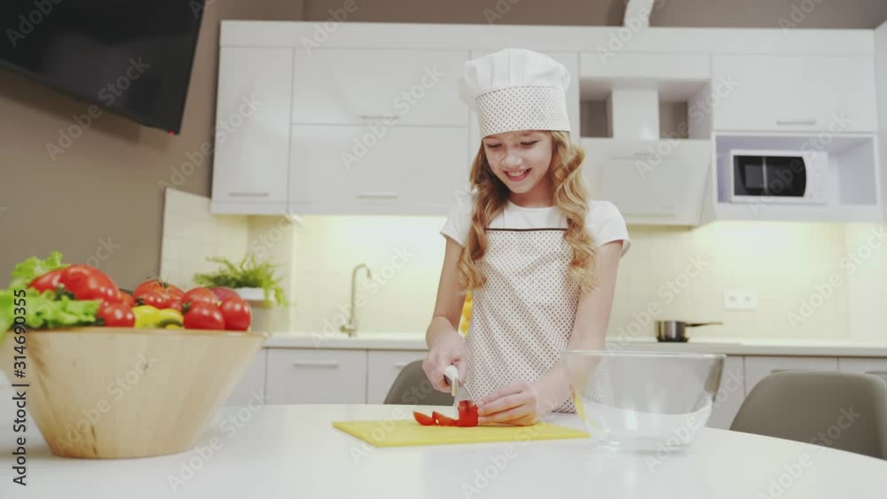Cute girl in kitchen apron and hat cutting tomato for salad Cute girl in kitchen apron and hat cutting tomato for salad