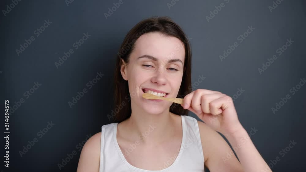 Close up of cute and pretty brunette woman brushes teeth wooden toothbrush, wears white t-shirt, actively works through all parts of her mouth hygiene on grey background. Dental care and reuse concept Close up of cute and pretty brunette woman brushes teeth wooden toothbrush, wears white t-shirt, actively works through all parts of her mouth hygiene on grey background. Dental care and reuse concept