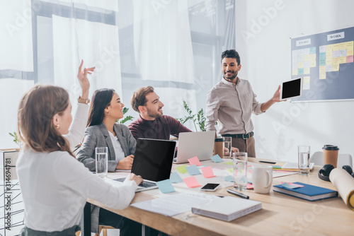bi-racial scrum master looking at businesswoman with raising hand near coworkers photo