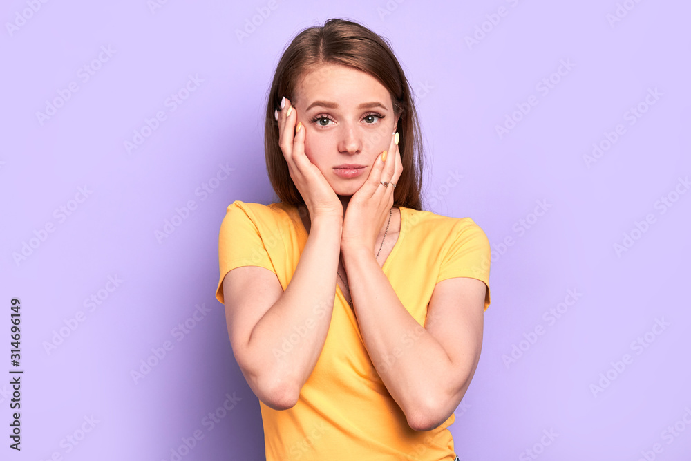 Stupefied sad girl keeps hands on her face, looks with upset expression in camera, has embarrassed and contemplative expression, received unpleasant news. People emotions concept.