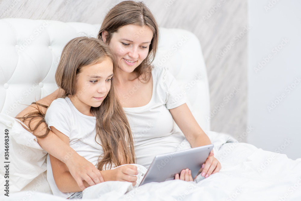 Young mother and her little daughter are using a digital tablet and smiling while sitting on the bed at home Young mother and her little daughter are using a digital tablet and smiling while sitting on the bed at home
