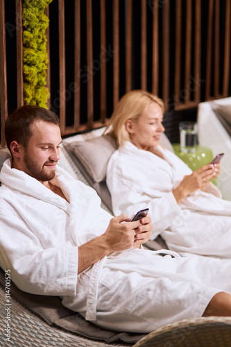 Massage and body care, blonde woman in white bathrobe having rest in comfortable chair together with handsome boyfriend, resting after aromatherapy massage, holding cell phones, sitting in silence