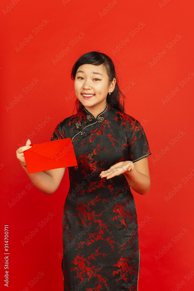 Chinese New Year 2020. Asian young girl's portrait isolated on red background. Female model in traditional clothes looks happy, smiling and showing red envelope. Celebration, holiday, emotions.