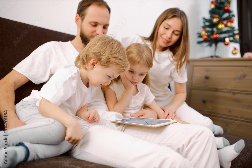 beautiful family watch video on modern tablet sitting on sofa together, dressed in white casual clothes, look at screen of tablet