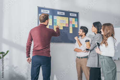 back view of scrum master pointing with finger at board with letters near multicultural coworkers