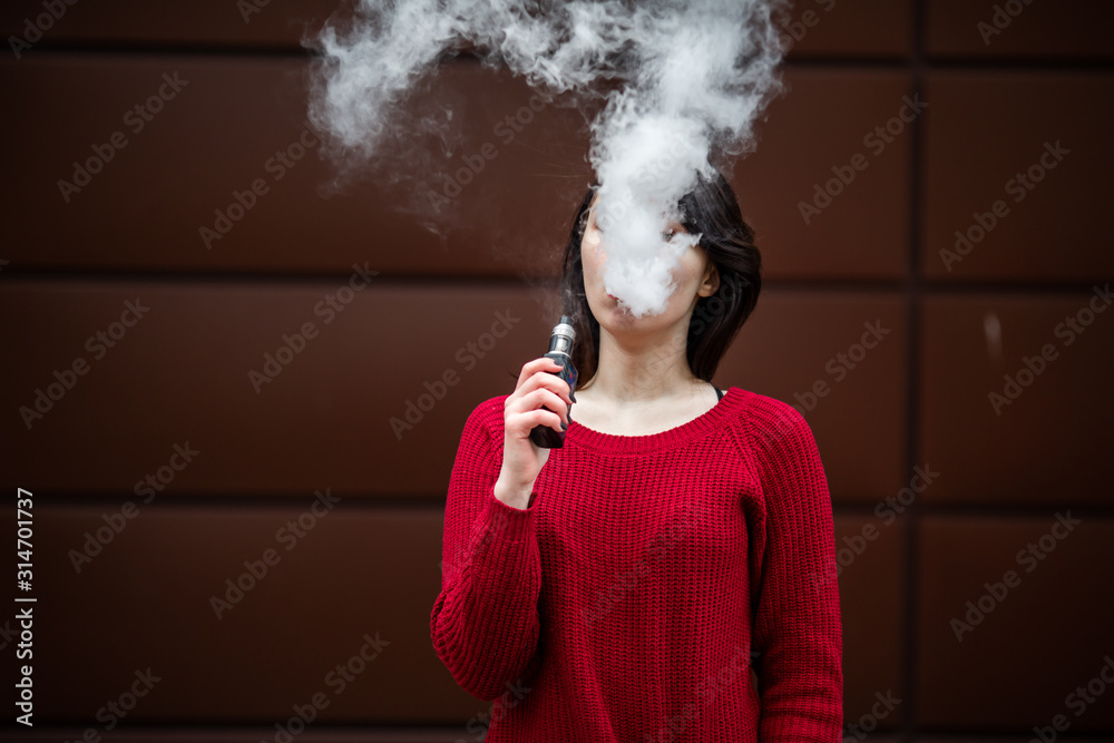 Vape teenager. Young pretty white caucasian girl in a red sweater smoking an electronic cigarette opposite modern brown background on the street in the winter. Deadly bad habit. - obrazy, fototapety, plakaty 