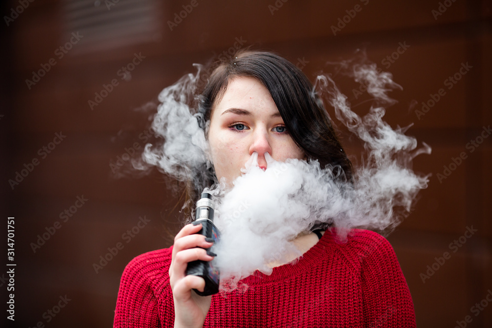 Vape teenager. Young pretty white caucasian girl in a red sweater smoking an electronic cigarette opposite modern brown background on the street in the winter. Deadly bad habit.