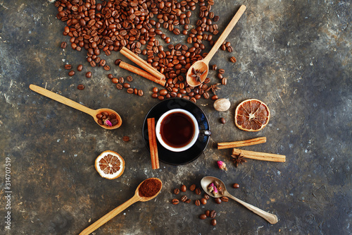 Cup of hot aromatic turkish coffee with a cinnamon stick on a saucer. Spoons with spices, dried roses, coffee beans and ingredients for making coffee are round the cup, flat lay. Dark aged surface